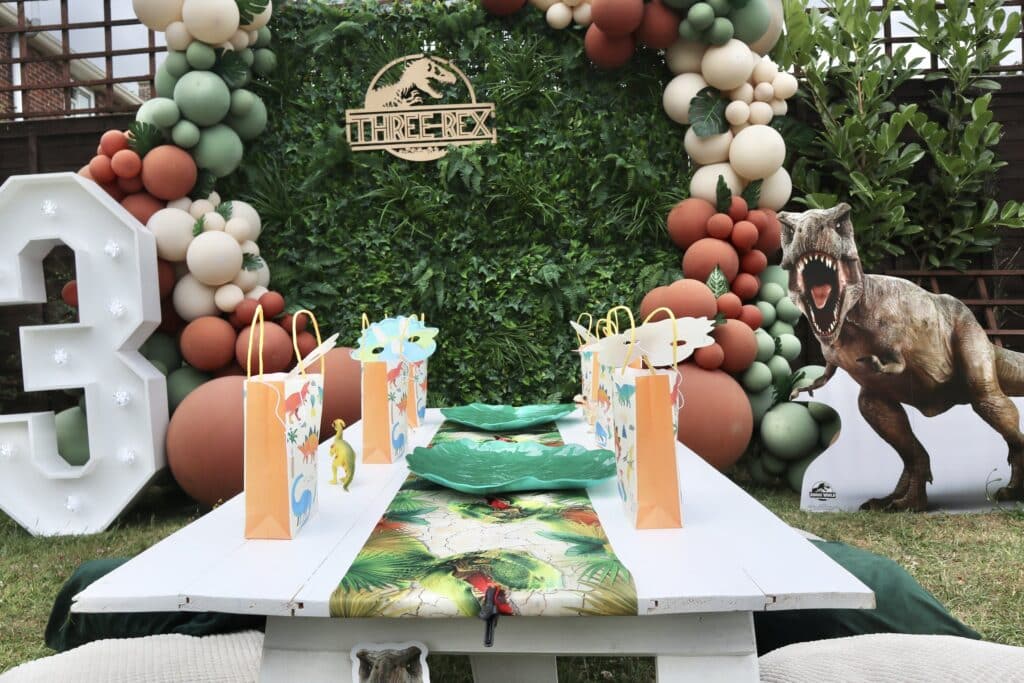 Backyard Three-Rex Dinosaur Party Decorations with white wooden table setup, T-Rex cardboard cutouts and balloon arch